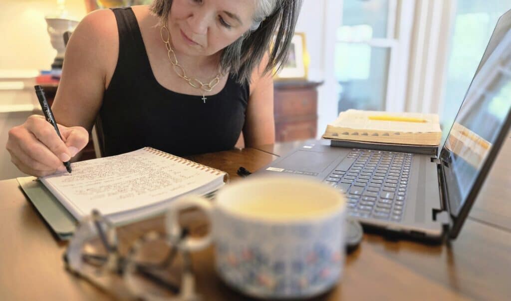 a lady writing with a coffee mug, glasses and computer also in the photo