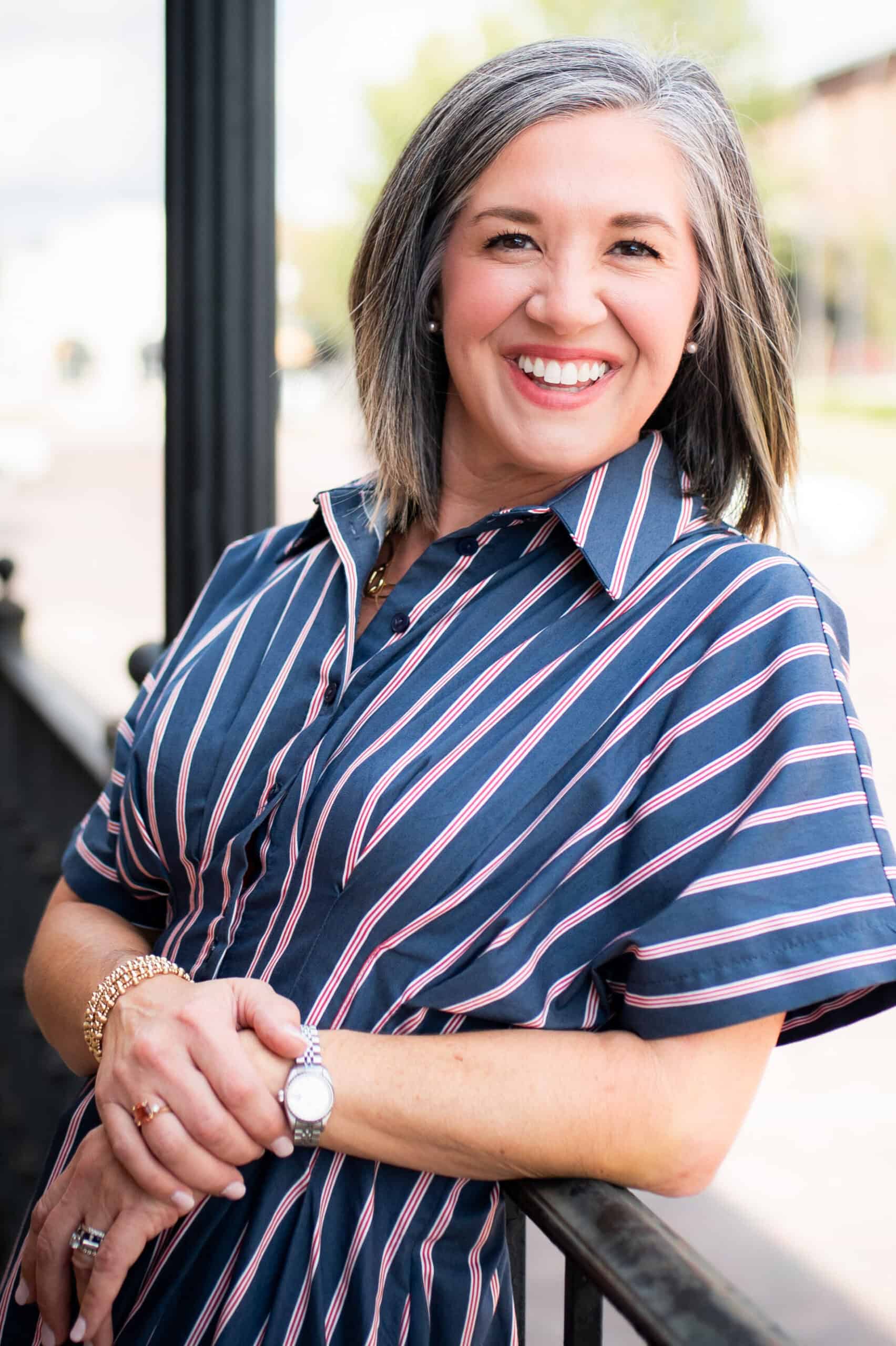 A photo of the author, in a blue striped dress, leaning on a railing. She is smiling and happy.